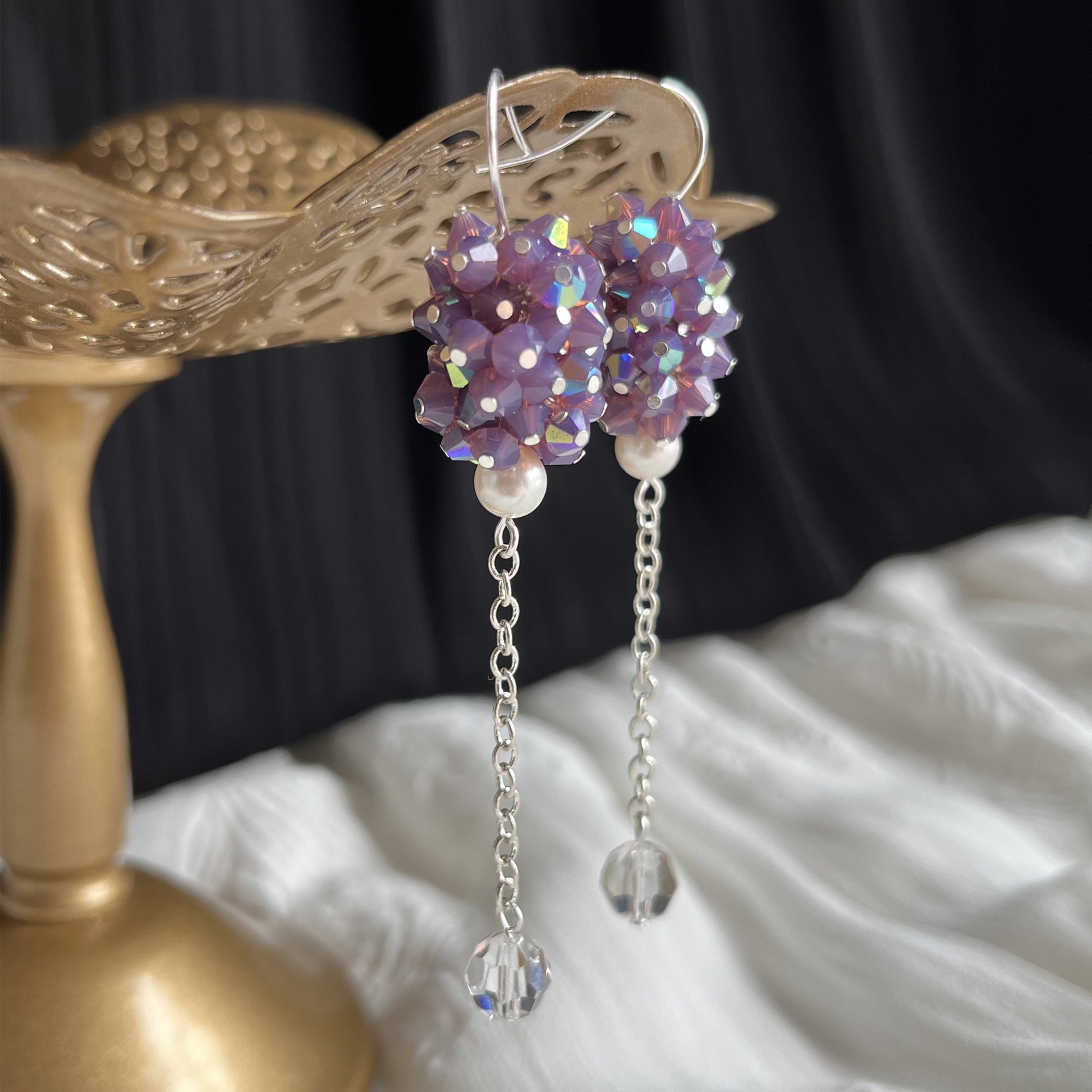Close-up side view of purple crystal and pearl earrings on a brass stand, showing sparkle and depth.